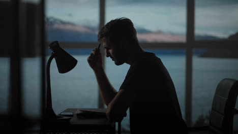 A-thoughtful-man-sitting-at-a-table-with-a-desk-lamp-writes-with-a-pen.-Dark-time-of-the-day-twilight-on-the-background-of-the-window-a-sad-man-thinks-and-writes